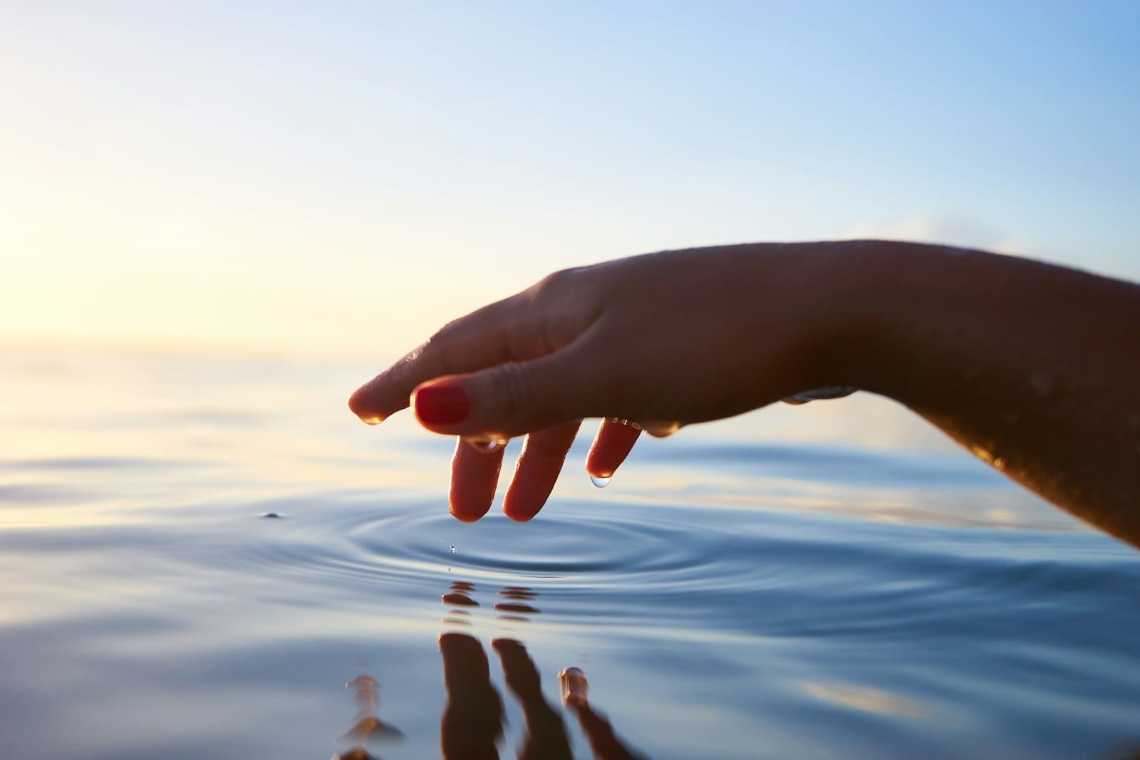 Resilient-Now: A hand with red nail polish hovers just above calm water, creating gentle ripples under a clear sky with soft sunlight.