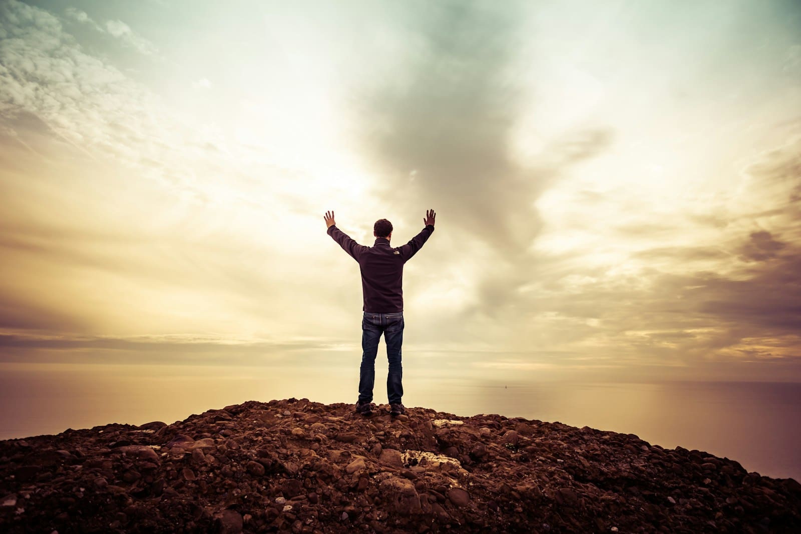 Resilient-Now: A person stands on a rocky hilltop with arms raised, facing a cloudy sky and a distant horizon at sunset or sunrise.