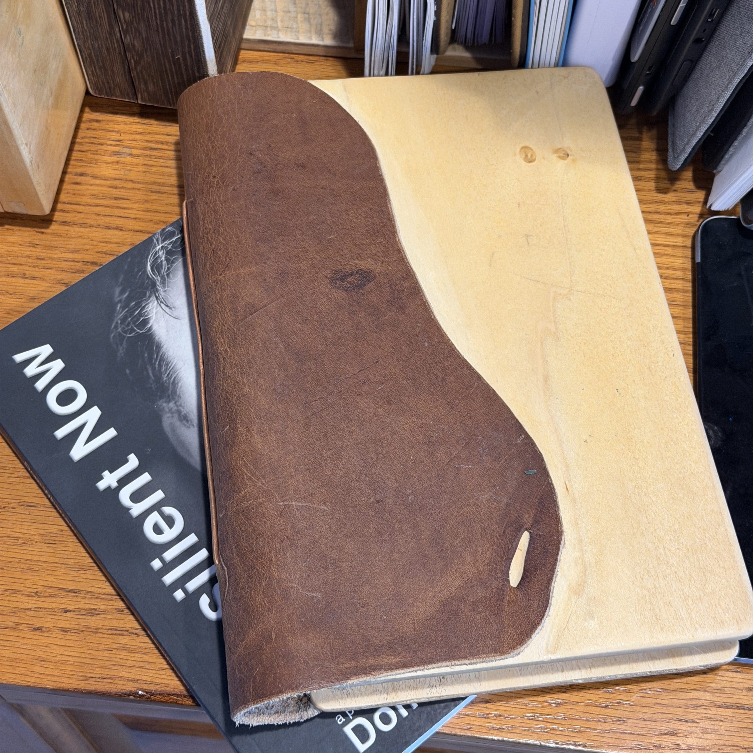 Resilient-Now: A wooden notebook with a partial brown leather cover rests on top of a book titled "Silent Now" on a wooden desk.
